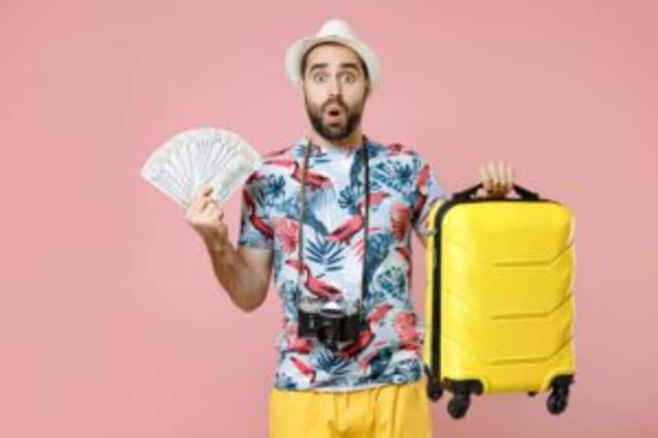 Homem com expressão surpresa em fundo rosa segurando vários notas de dinheiro em uma mão e uma mala de viagem na outra