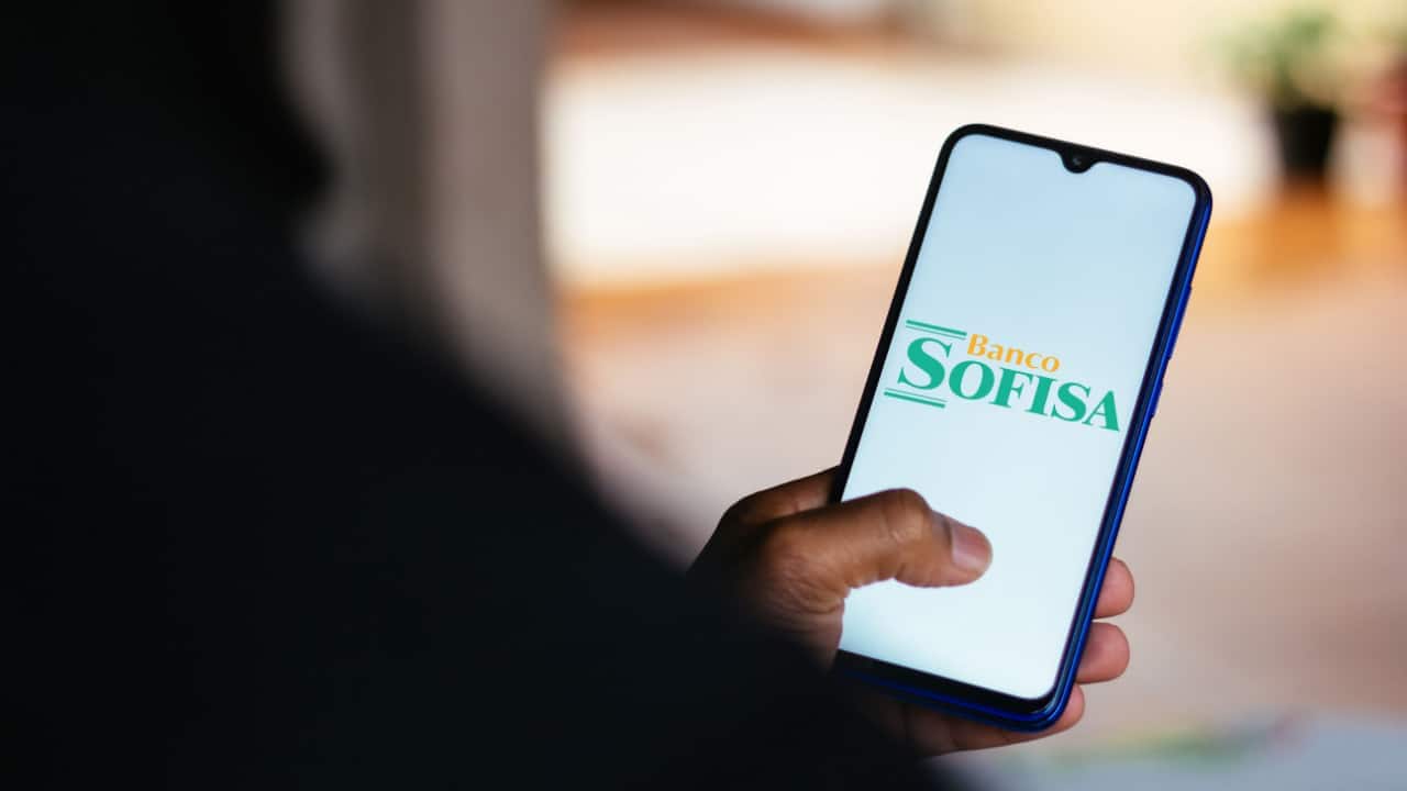 Pessoa de costas segurando celular que mostra logo do banco Sofisa Direto