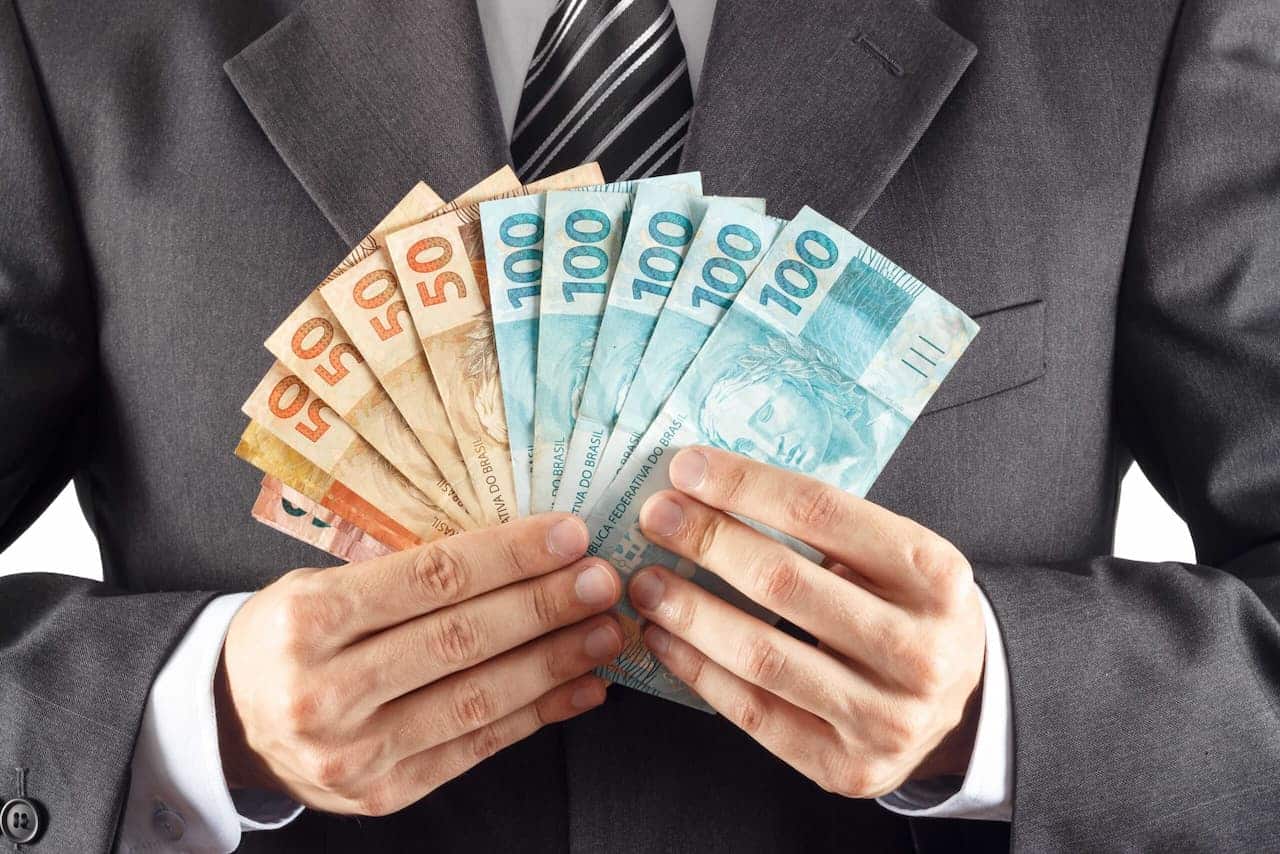 mãos de homem de terno segurando notas de R$ 100, R$ 50, R$ 20 e R$ 10