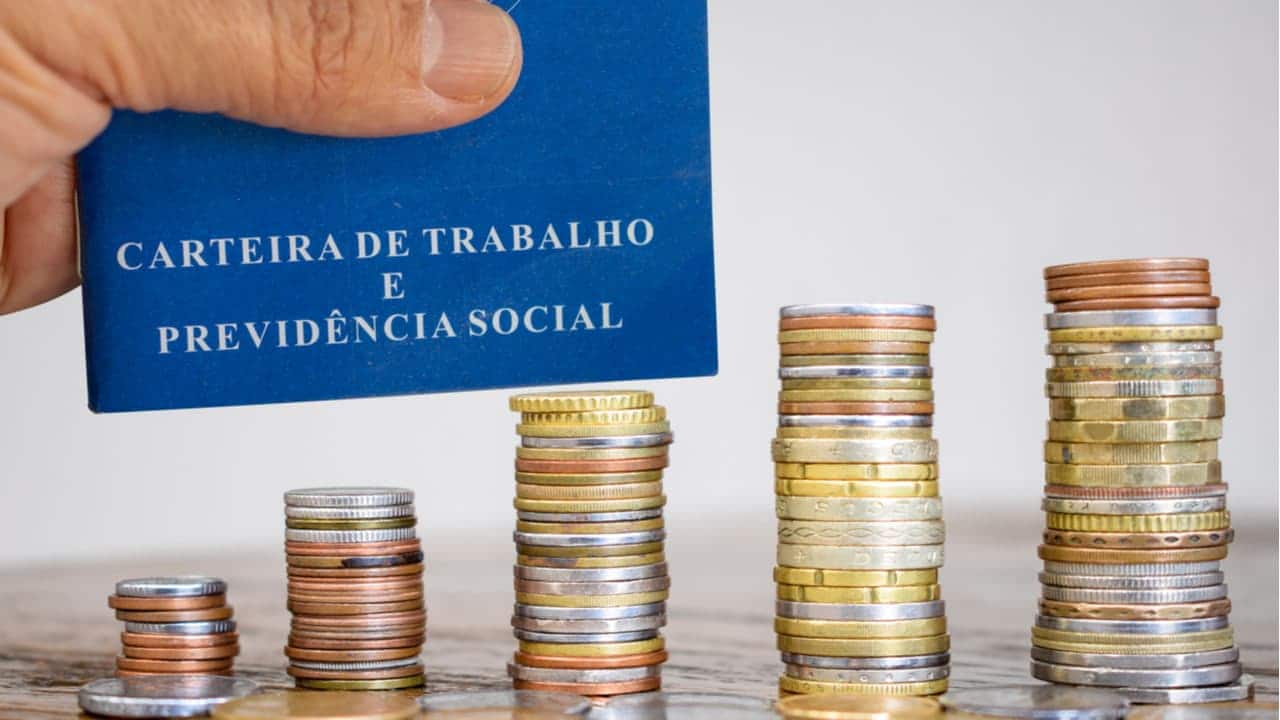Mão segurando Carteira de Trabalho acima de pilhas de moedas que aumentam de tamanho da esquerda para a direita