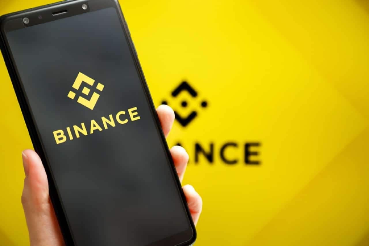 Imagem da logo do Binance na tela de um celular e, ao fundo, uma tela com a logo novamente.