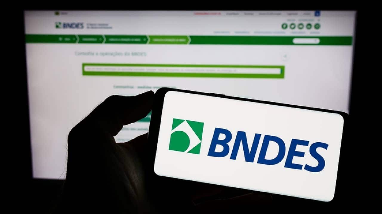 Imagem de uma pessoa segurando celular com a tela na logo do BNDES.