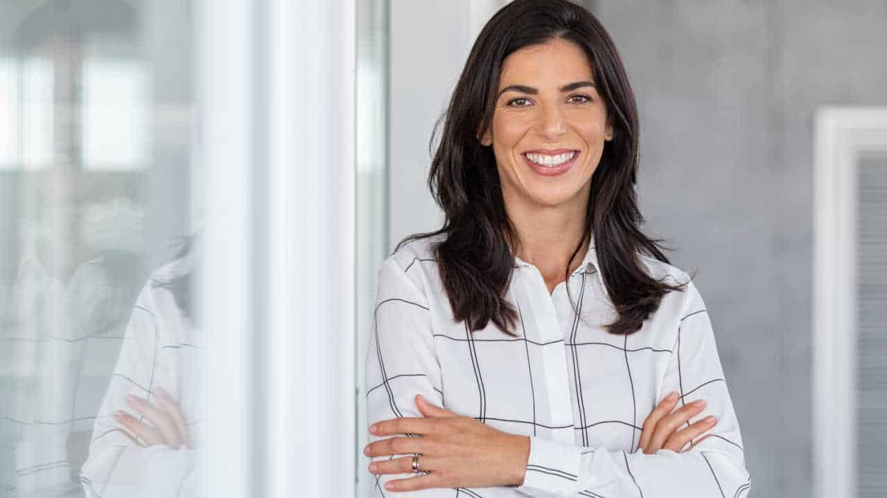 Mulher de cabelos pretos abaixo dos ombros sorrindo enquanto está de braços cruzados. Usa uma camisa branca com listras pretas.