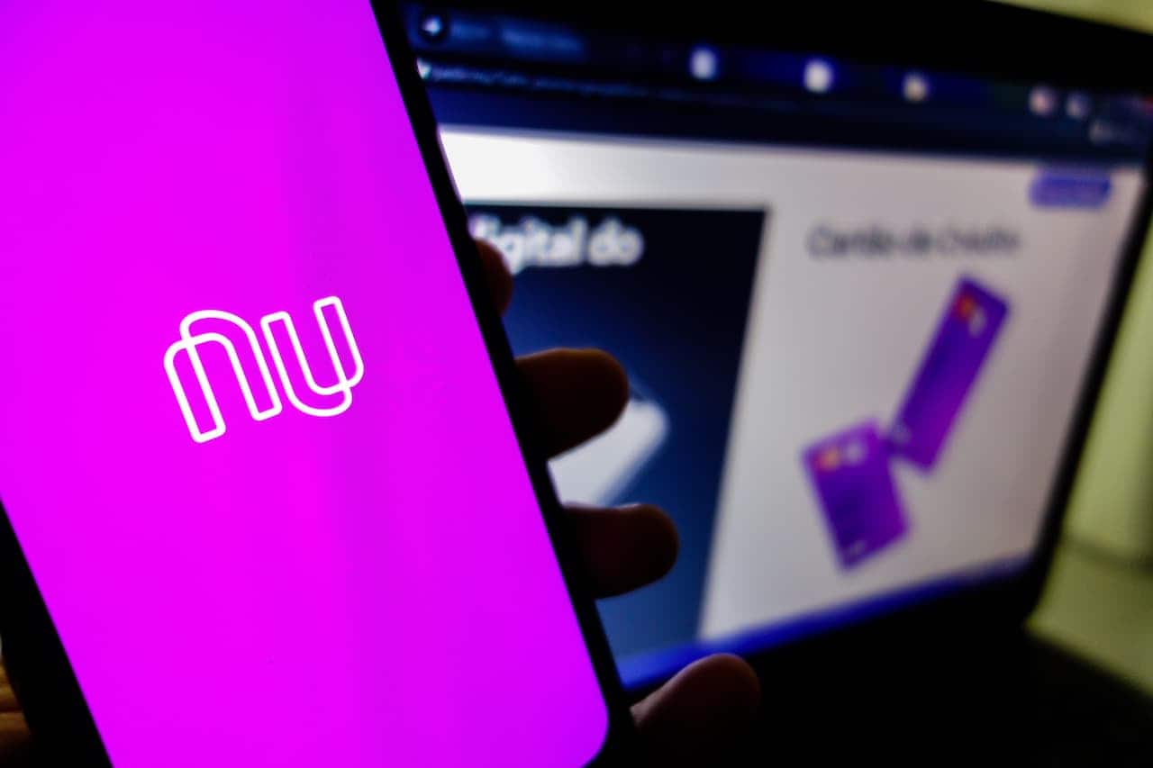 Celular com aplicativo Nubank e ao fundo uma notebook no site do Nubank