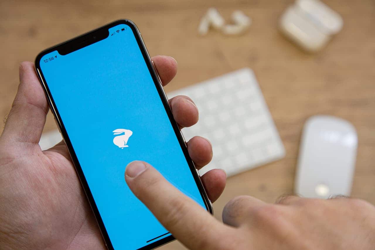 Smartphone aberto no aplicativo da Loggi (tela azul com um coelho). Ao fundo, há objetos como teclado e fone de ouvido, mas fora de foco