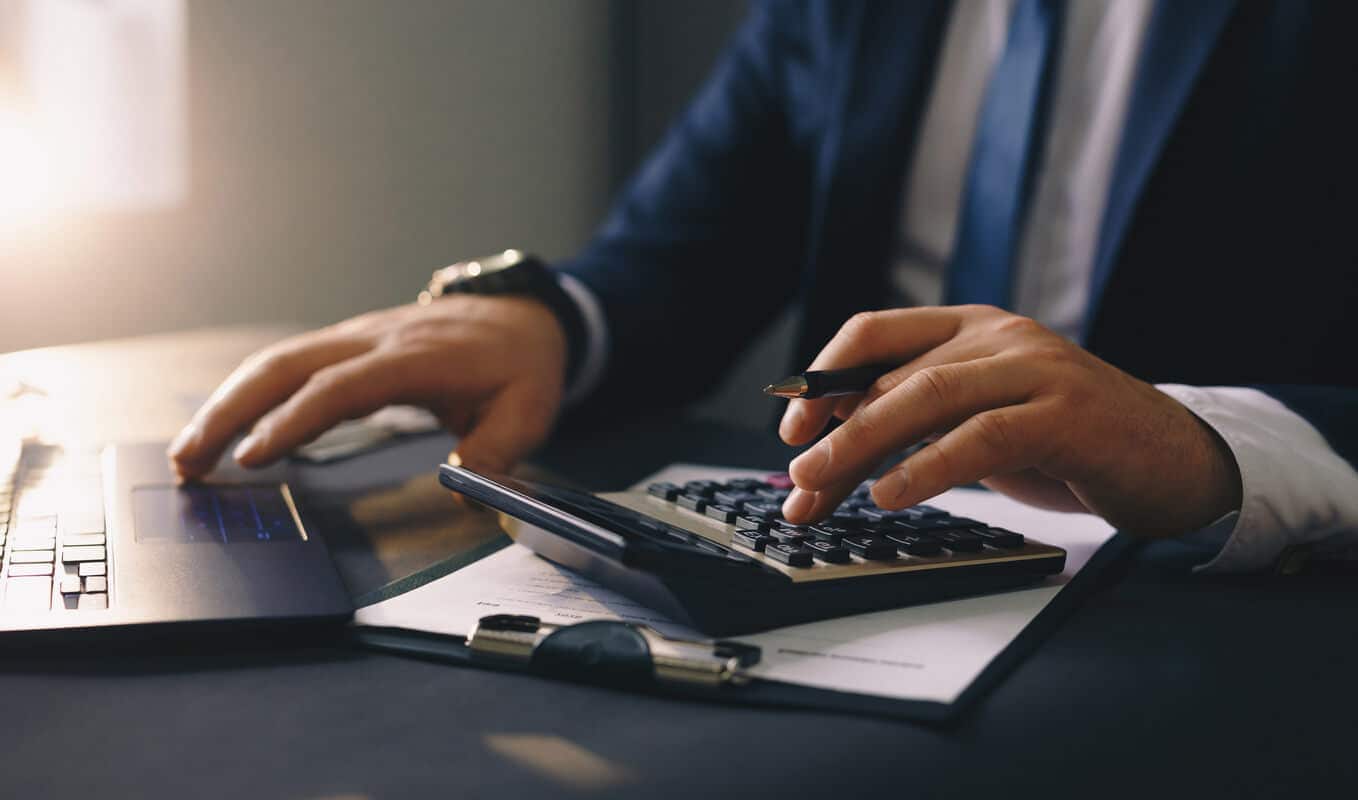 Na foto há um notebook e mãos de um homem de terno usando uma calculadora
