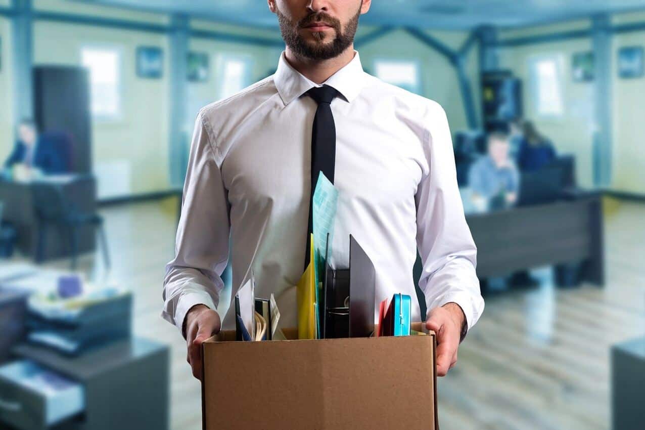 Imagem mostra homem segurando uma caixa de papelão com objetos de trabalho após ser demitido.