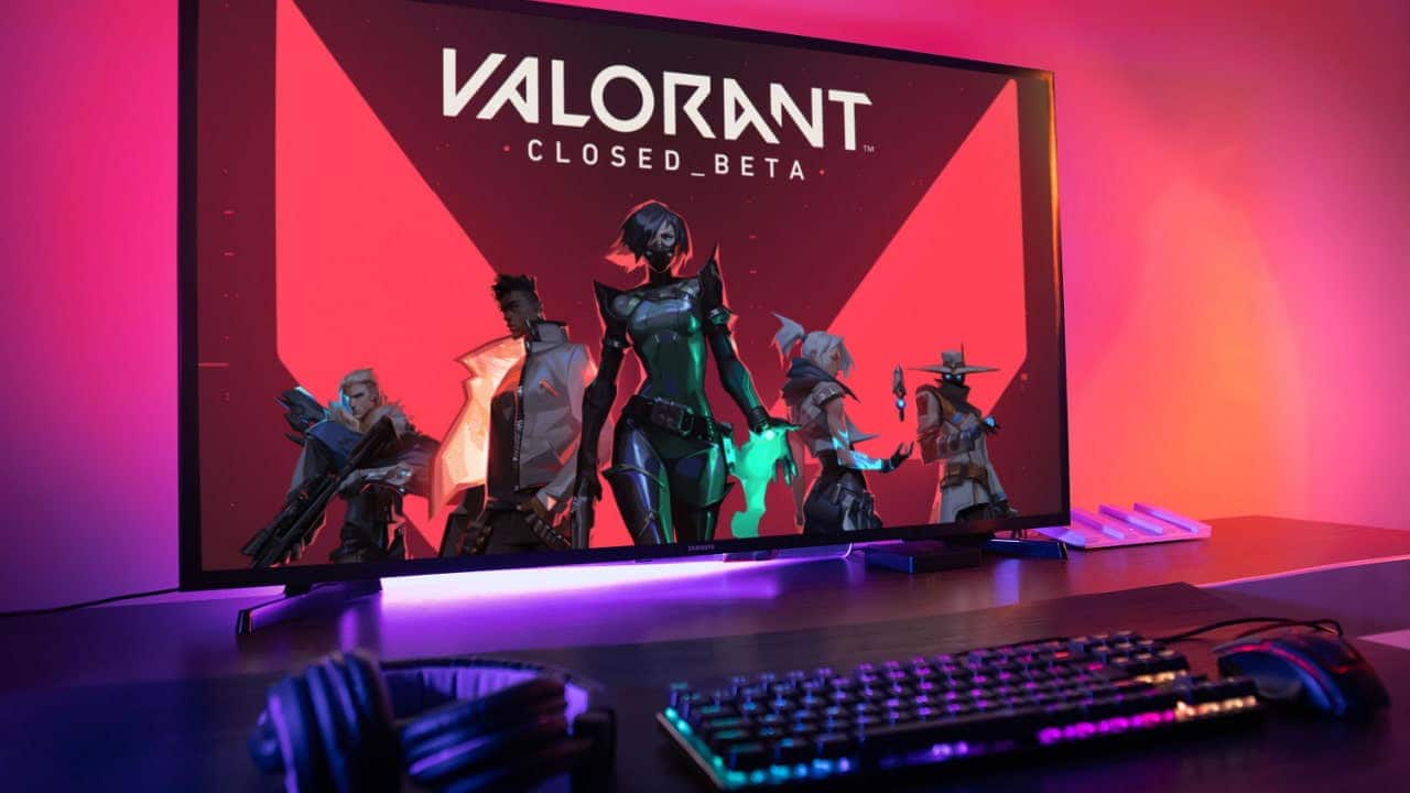 Monitor de um computador exibindo uma imagem de divulgação com personagens do jogo eletrônico Valorant. Sobre a mesma mesa, um teclado e fones de ouvido.