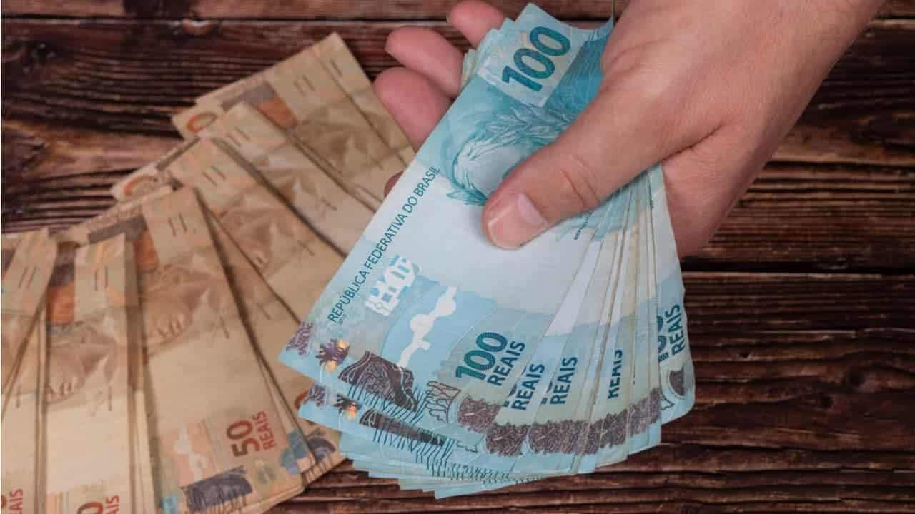 Mão segurando um leque de notas de cem reais. Abaixo, sobre uma superfície de madeira, várias notas de cinquenta reais