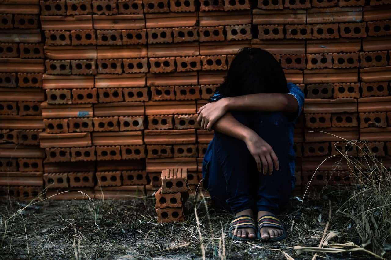 imagem de uma criança sentada no chão com a cabeça entre as pernas, expressando tristeza e atrás de vários tijolos empilhados