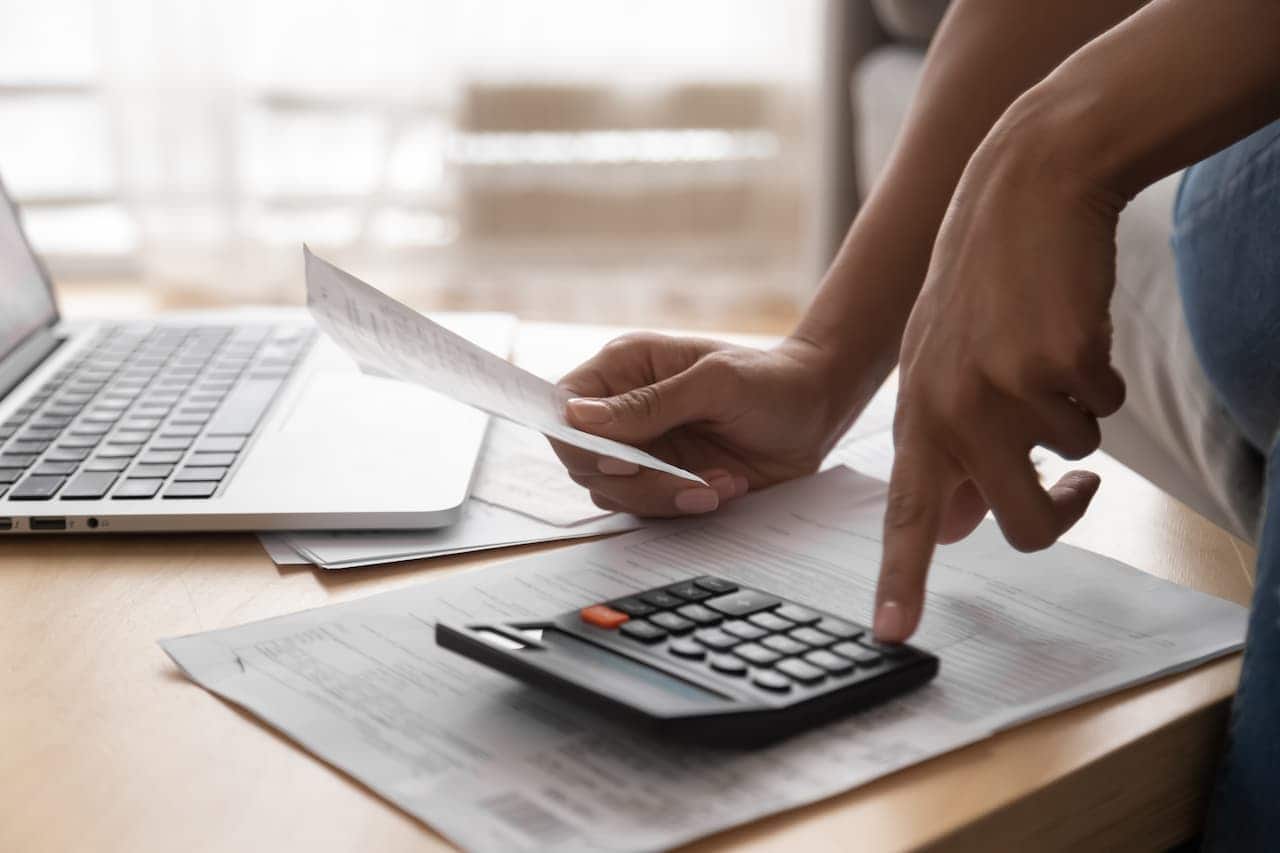 Mão segurando dívida enquanto disca em calculadora em cima da mesa onde também tem um laptop.