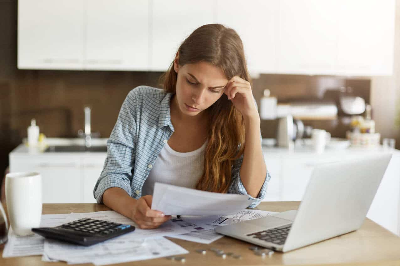 Na imagem há uma mulher jovem com alguns papéis e um laptop, verificando seu orçamento.