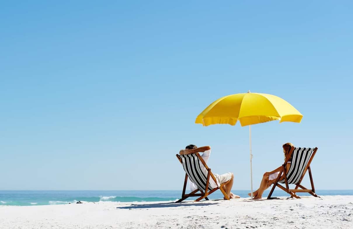 Duas pessoas aproveitando uma praia em frente ao mar, guarda sol e cadeiras