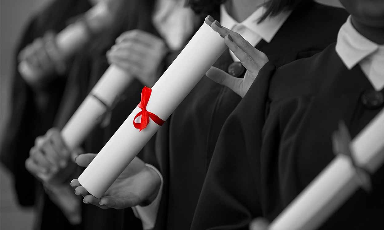 Imagem em preto e branco de estudantes vestidos com becas e segurando diplomas. O único elemento da imagem que está colorido é uma fita vermelha ao redor do diploma segurado por um aluno.