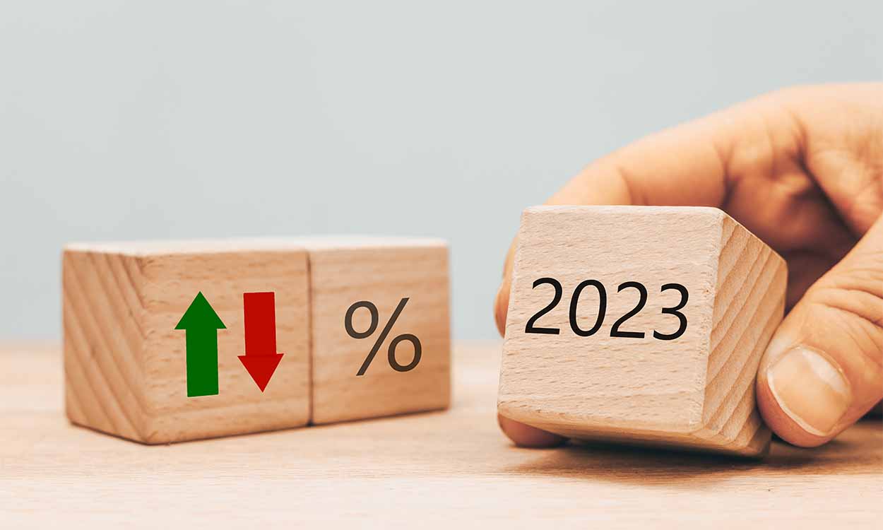 Cubos de madeira apoiados em superfície com setas verde e vermelha em sentido contrário. Ao lado, outro cubo com sinal de % e uma mão segurando outro cubo escrito 2023.