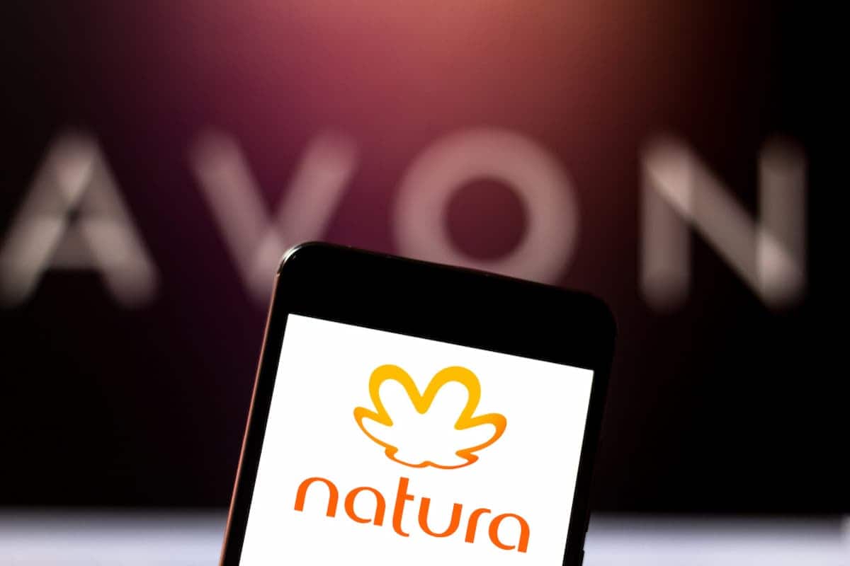 Logo da Avon ao fundo e um celular a frente com o logo da Natura