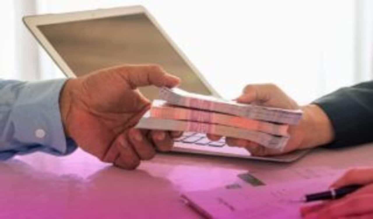 Uma pessoa esticando a mão com três maços de notas de dinheiro para outra. A superfície em que estão é cor de rosa e há um notebook ao