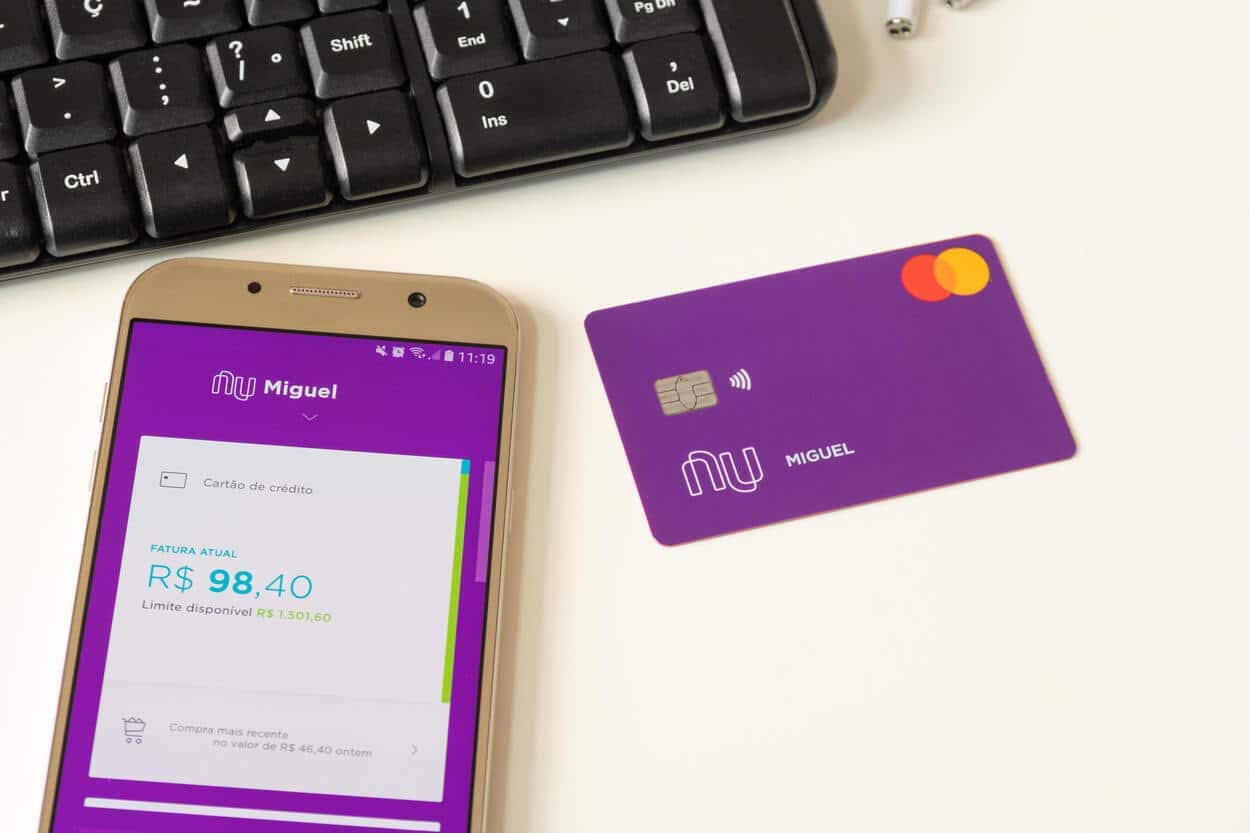 fundo branco, com teclado, celular com o app do Nubank em sua tela e o cartão físico ao lado.