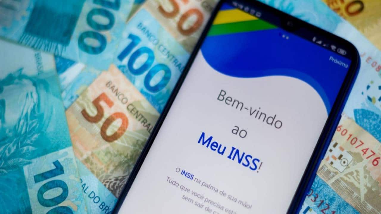 Celular mostrando o aplicativo Meu INSS sobre notas de cem e cinquenta reais