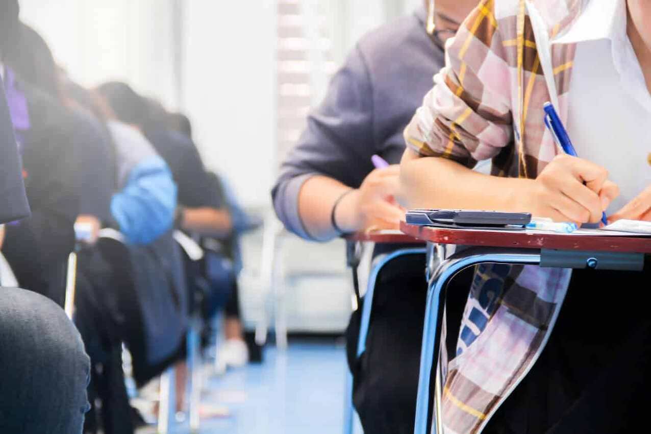 Pessoas sentadas em carteiras escolares, enfileiradas em uma sala de aula e realizando alguma atividade em papel, semelhante a uma prova.