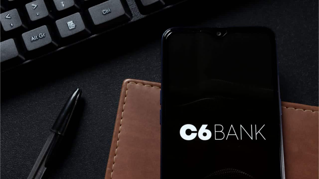 Ao lado de uma caneta e um teclado e posicionado em cima de uma carteira marrom, celular exibe logo do C6 Bank