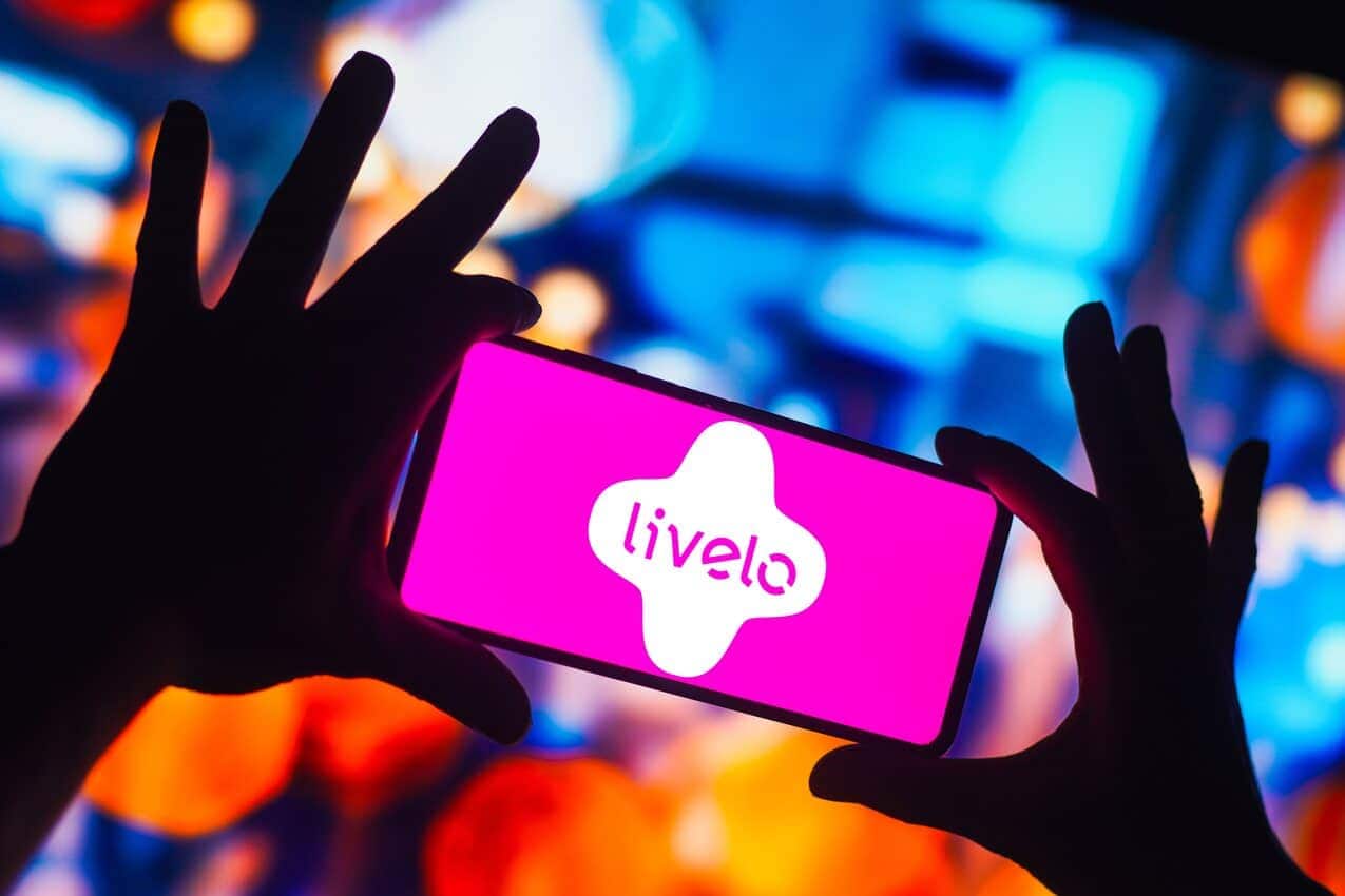 Mãos segurando celular com o logo da Livelo na tela, em um fundo desfocado colorido.
