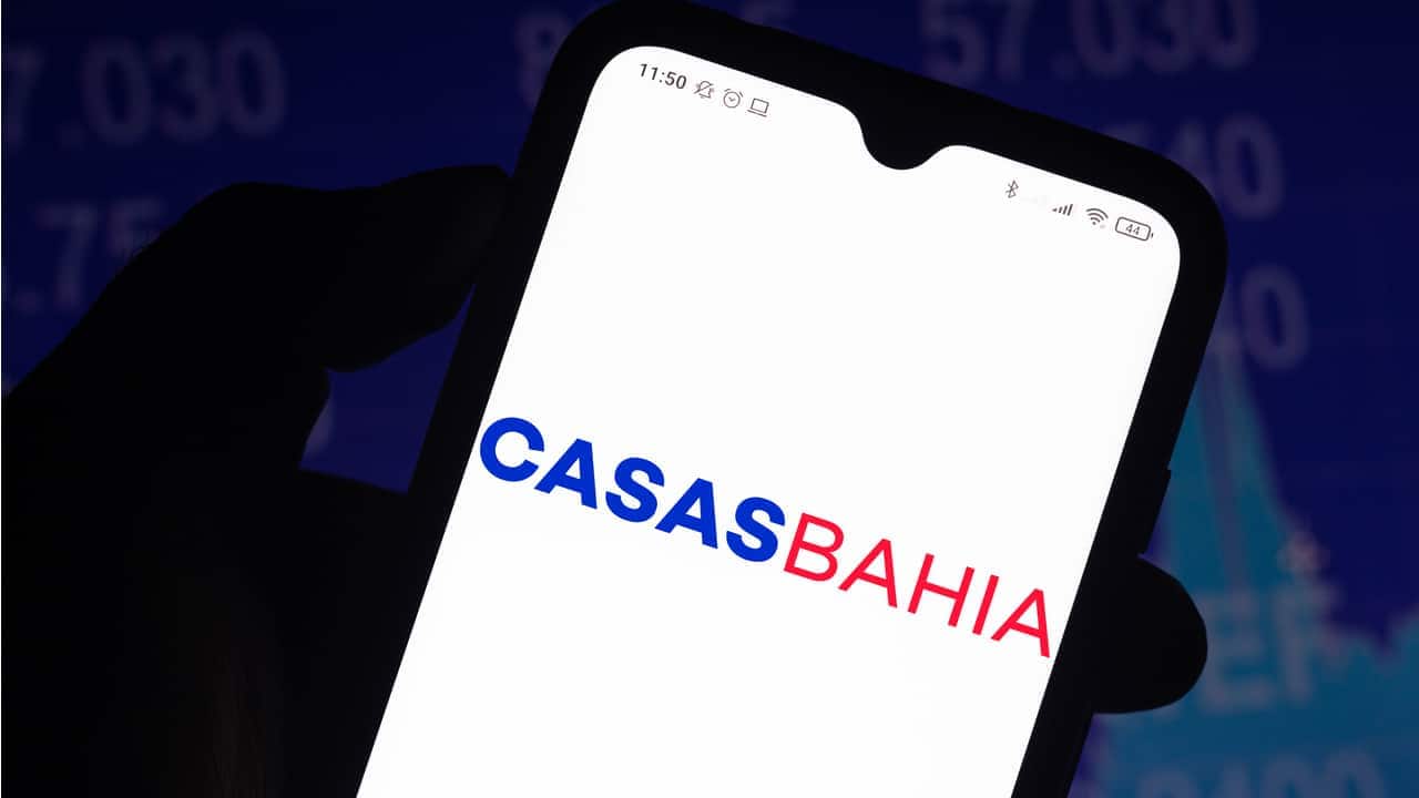 Mão em um ambiente escuro segurando um celular que exibe a logo da Casas Bahia