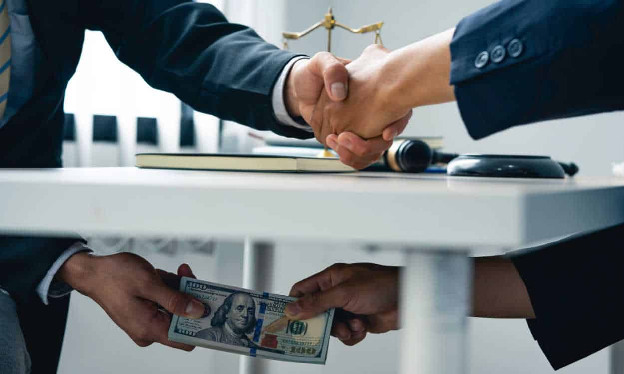Homens apertando as mãos enquanto trocam um maço de dinheiro por debaixo da mesa