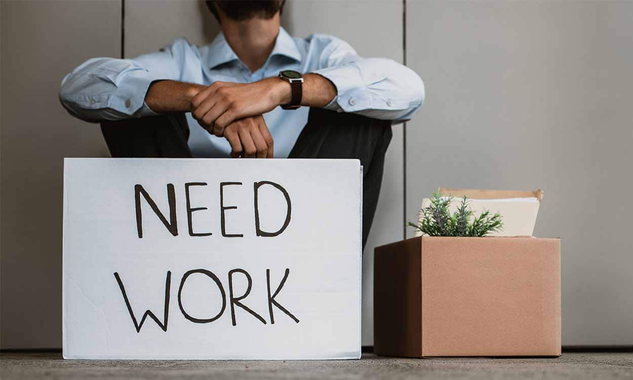 Homem sentado no chão, junto com uma caixa com todos os seus pertences do trabalho e um cartaz onde se lê "Precisa-se de trabalho"