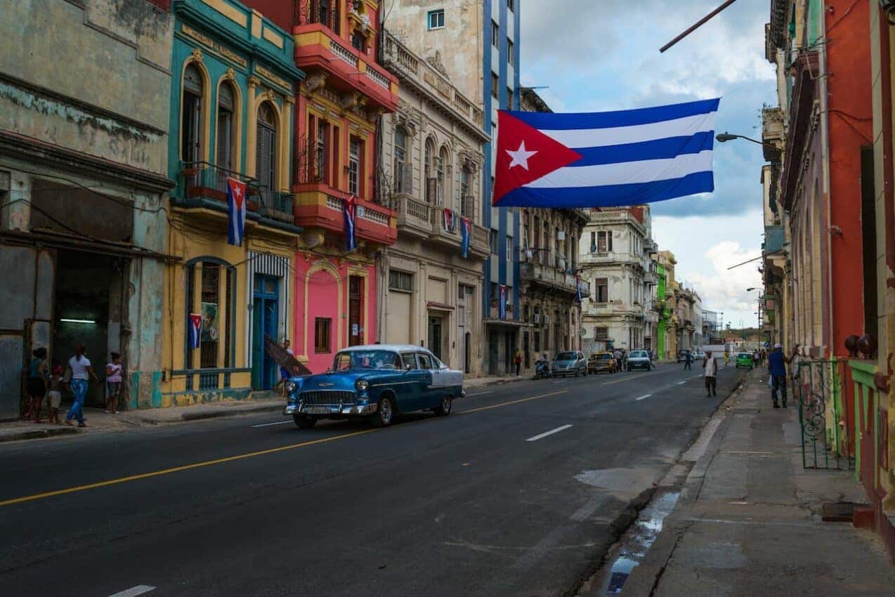 Imagem de Havana, em Cuba. Prédios coloridos e antigos, carro antigo e bandeira de Cuba hasteada