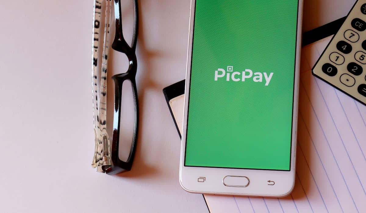 Aplicativo PicPay em celular sobre mesa, com óculos ao lado e folhas embaixo