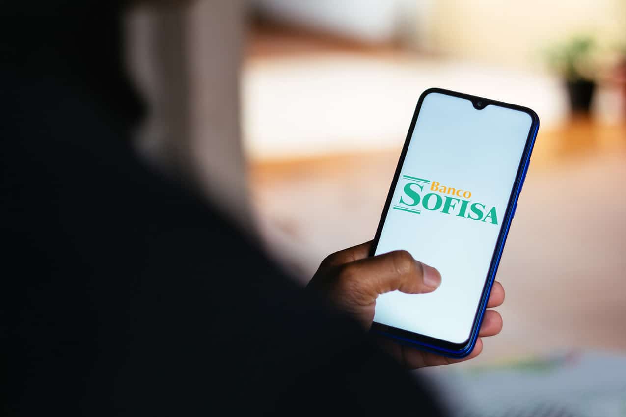 Pessoa de costas segurando celular que mostra logo do banco Sofisa Direto