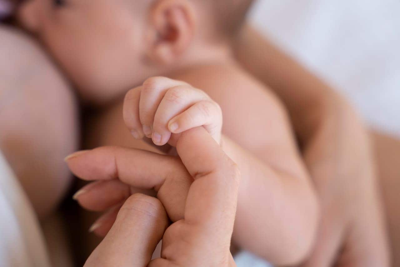 Imagem de um bebê sendo mamando e sua mãe segurando um de suas maõs. vacina
