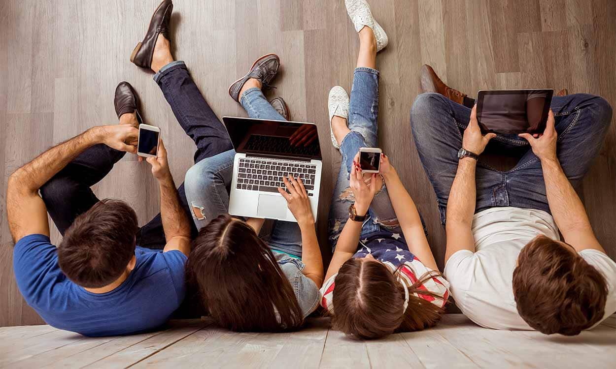 Na imagem adolescentes sentados no chão com aparelhos eletrônicos.