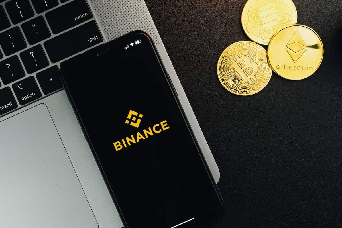 Aplicativo da Binance em celular sobre notebook, com tokens de criptomoedas ao lado