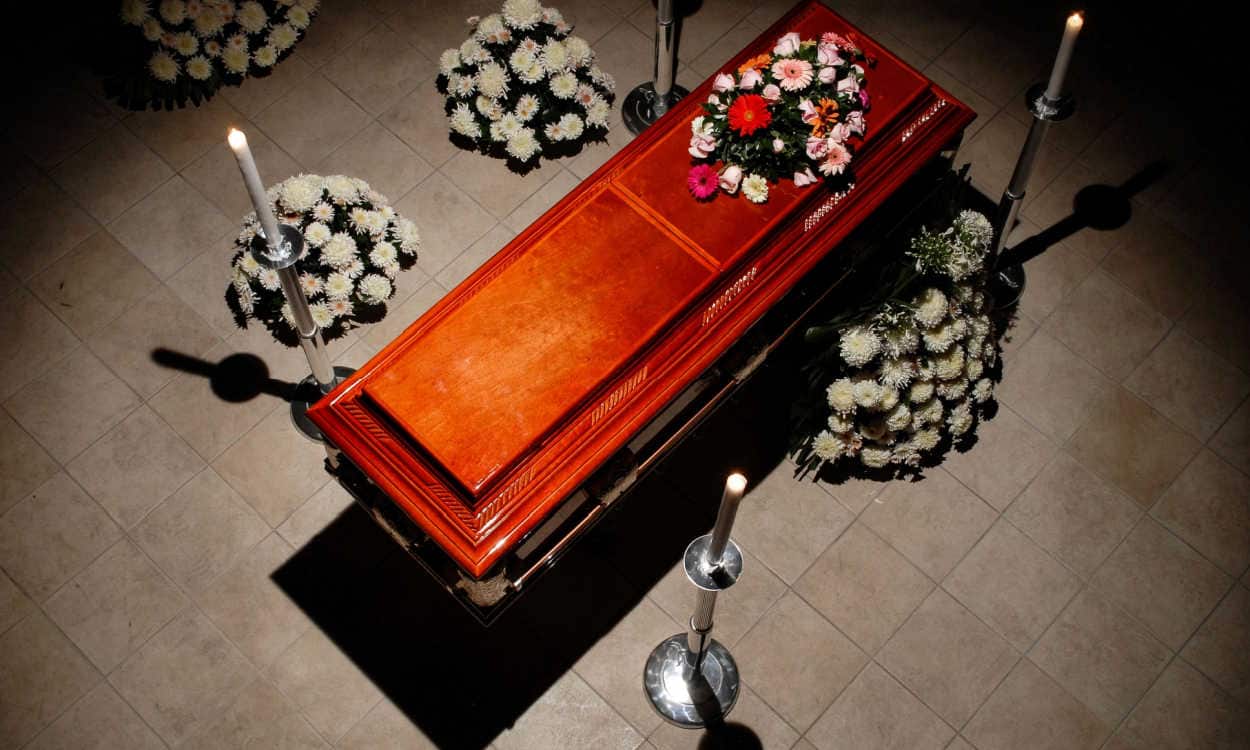 Caixão rodeado por flores e velas, em uma sala pouco iluminada