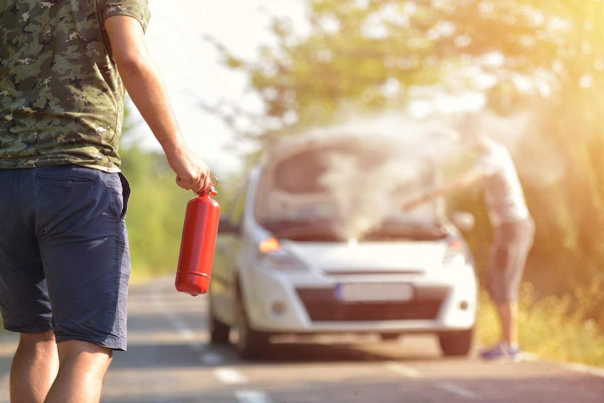 Homem com um extintor na mão corre para ajudar outra pessoa com o carro quebrado e soltando fumaça.