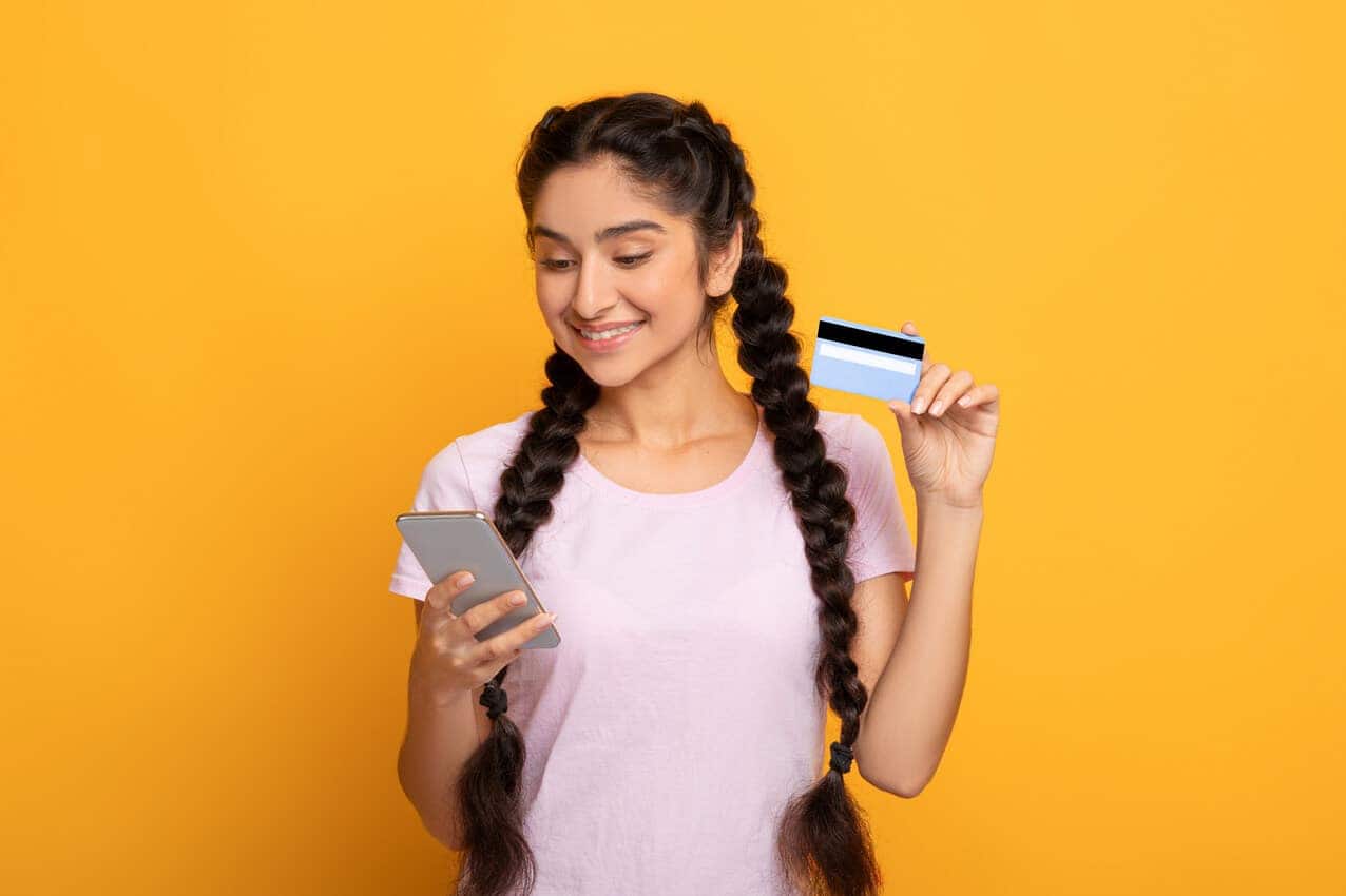 Mulher segurando cartão de crédito em uma das mãos e na outra um celular.