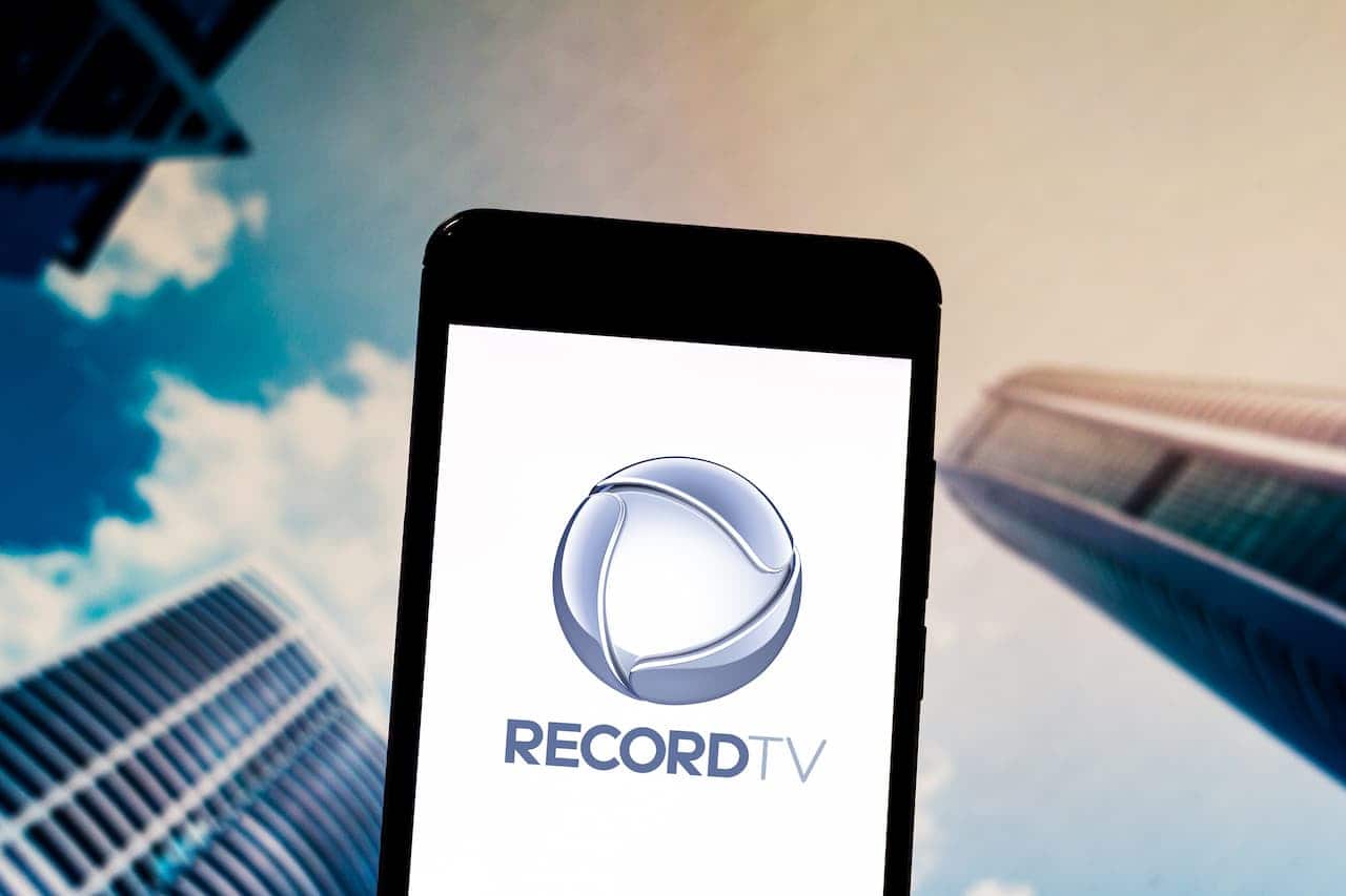 Celular exibindo a logo da Record TV, ao fundo tem alguns arranha-céus no meio de nuvens