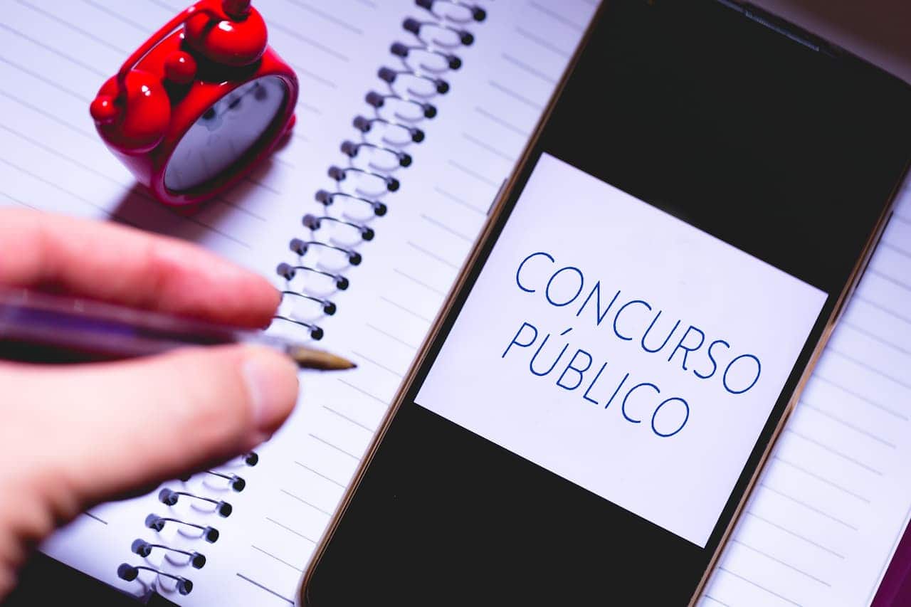 Pessoa com caneta na mão, se preparando para escrever em caderno com os dizeres "concurso público".