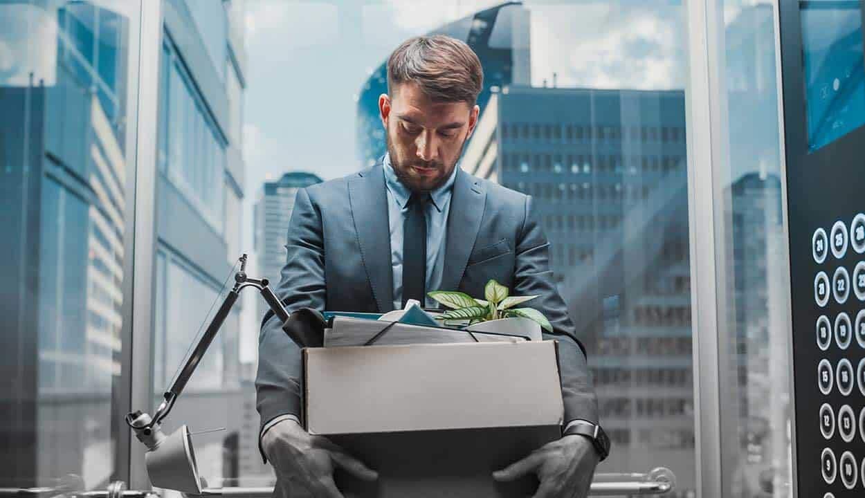 Homem de terno com caixa nas mãos e cabeça baixa. Prédios modernos ao fundo.