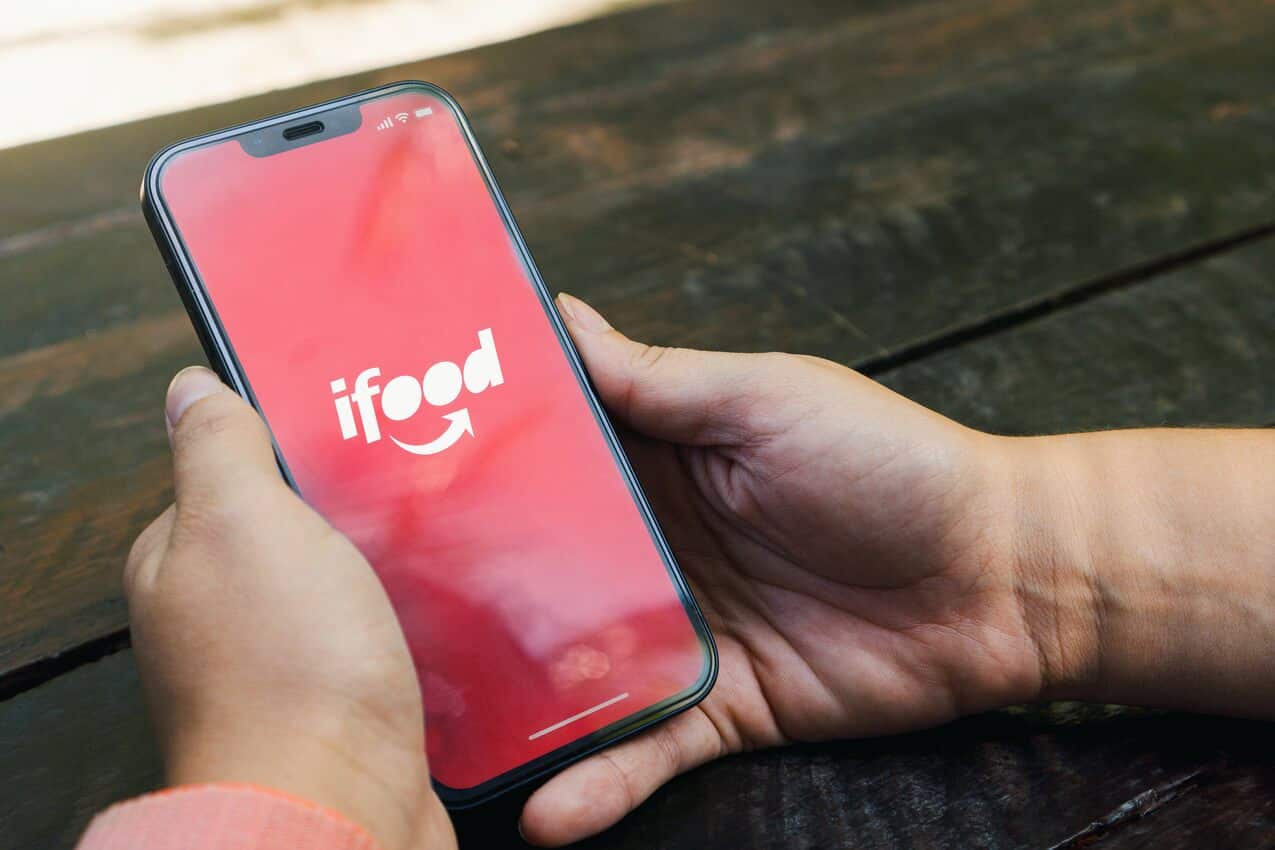 Na imagem, mãos segurando um celular com a tela inicial do iFood aberta.