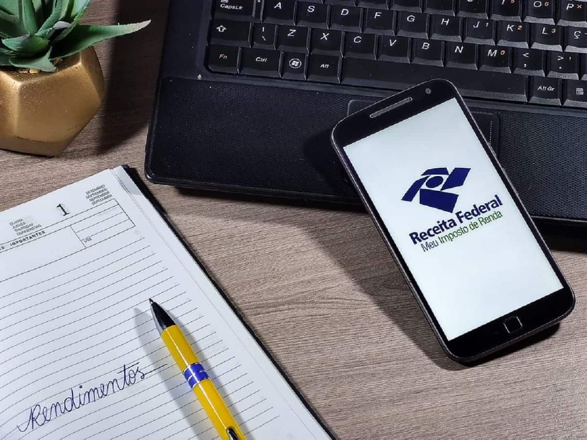Imagem de um celular com a tela na logo da Receita Federal Imposto de Renda e um caderno ao lado.