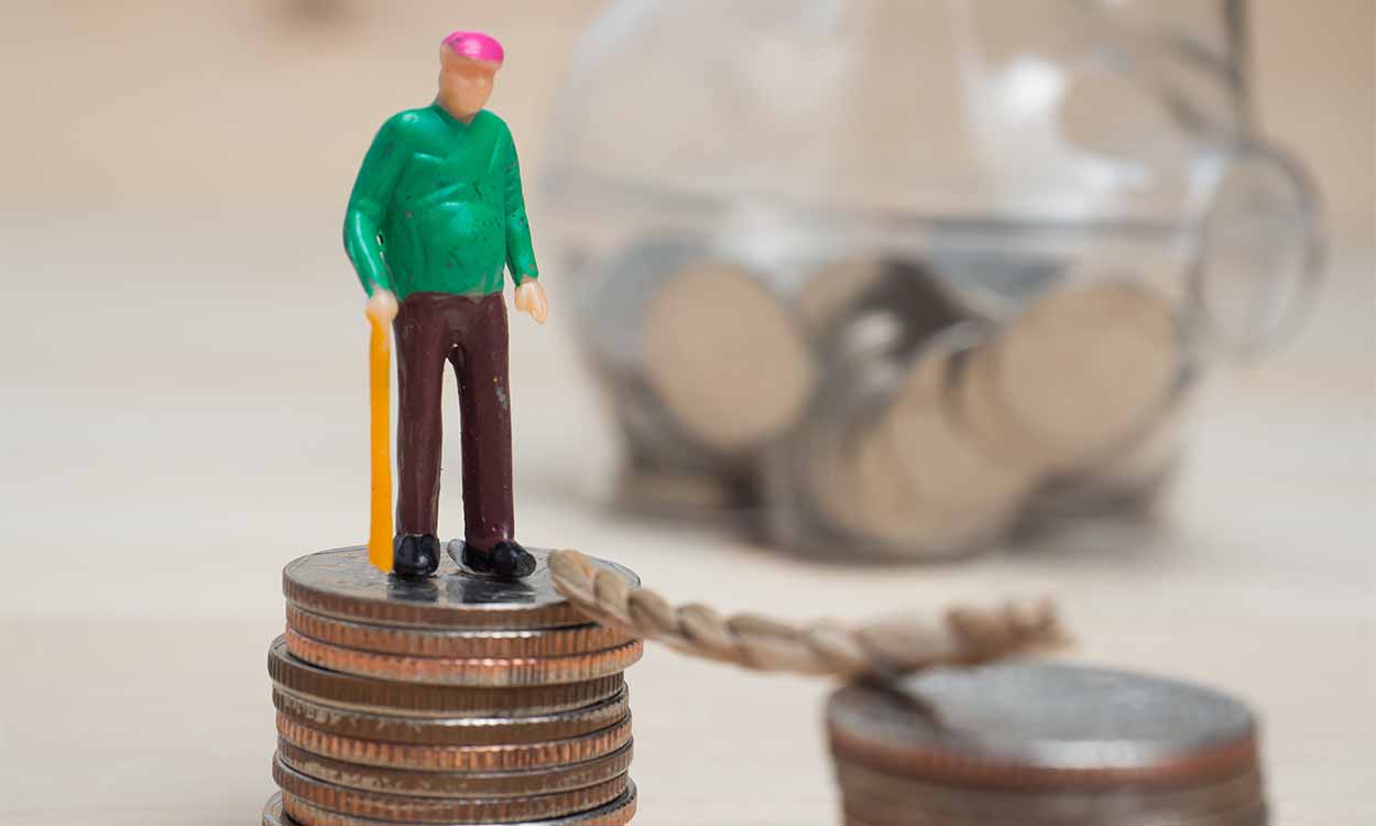 Na imagem, há uma um boneco em miniatura de um idoso. Ele está em cima de uma pilha de moedas. Essa pilha tem uma cordinha que leva a outra pilha igual, como uma ponte. Ao fundo, desfocado, um cofre de porquinho transparente e preenchido até a metade com moedas