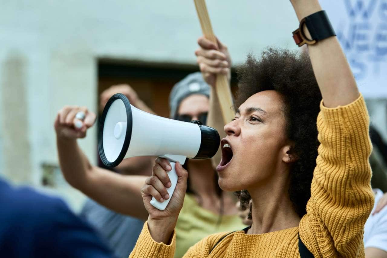 mulher protestando usando megafone, com outras pessoas ao fundo, todos de mãos erguidas