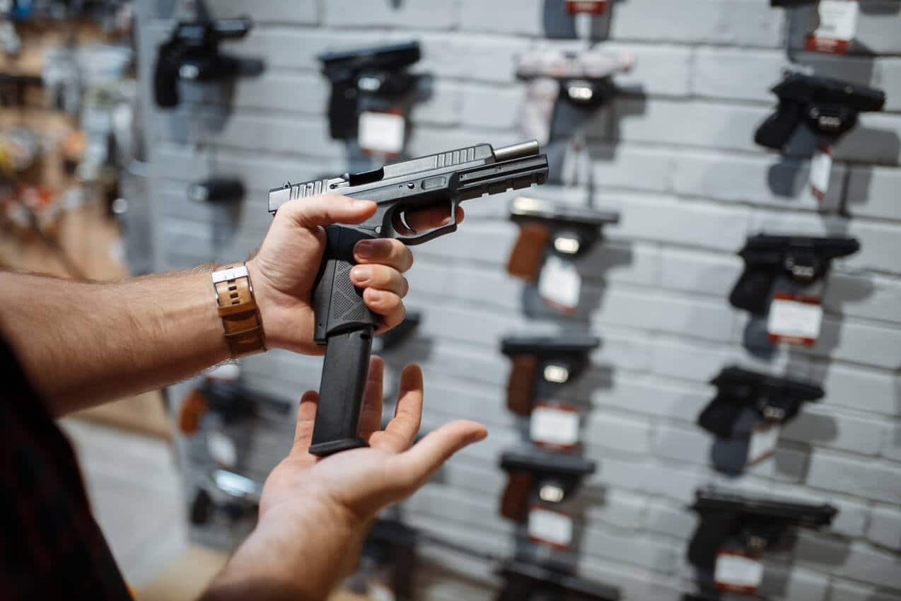 Pessoa segurando uma pistola, colocando o cartucho. Ao fundo, há outras armas parecidas em uma parede, penduradas, representando clube de tiro