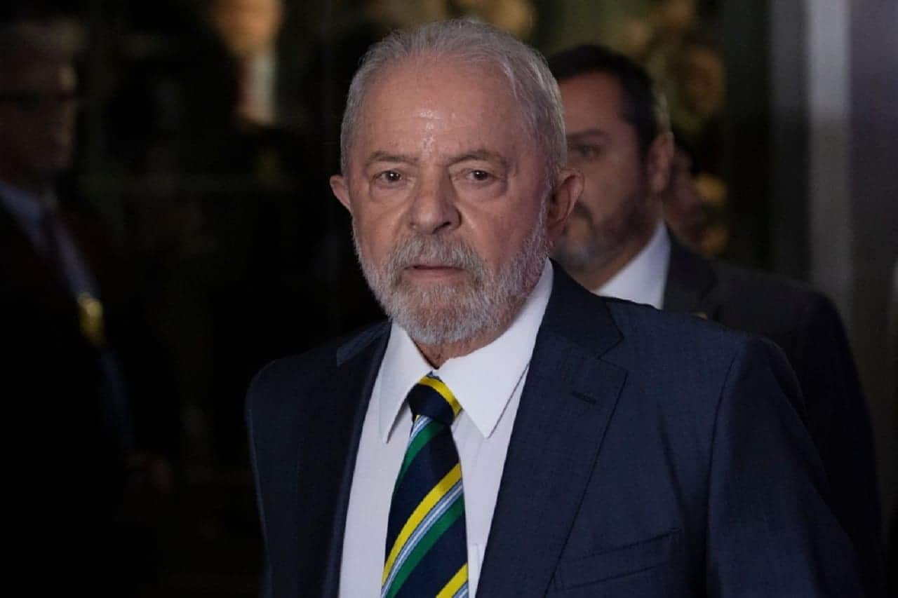 Presidente Lula com expressão séria. Ao fundo, há dois homens desfocados, um quase invisível