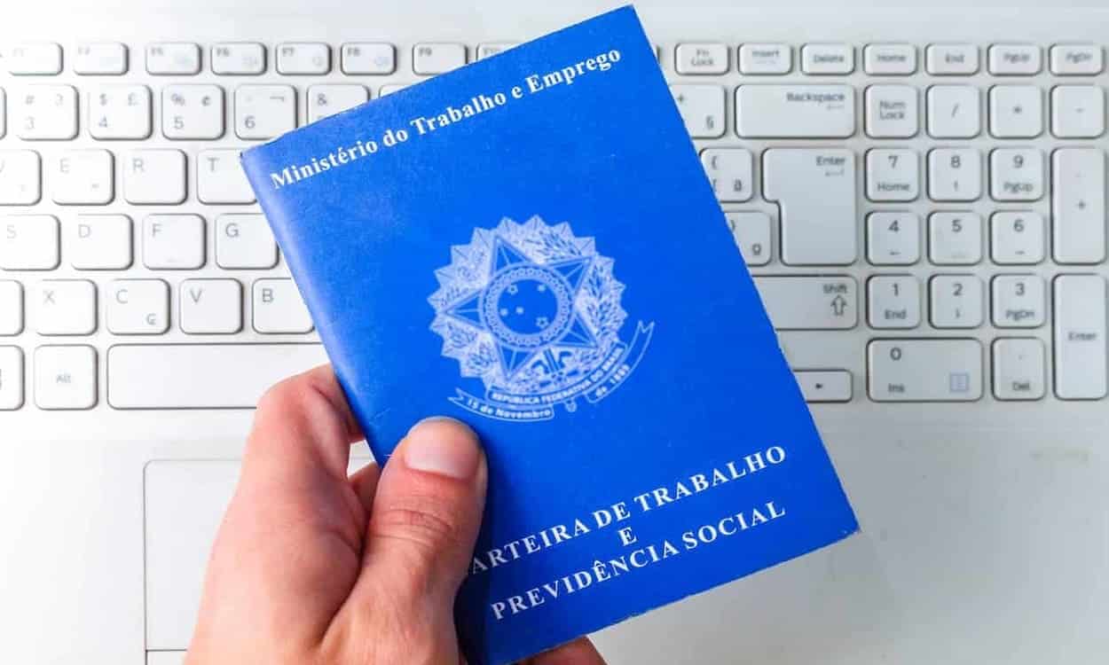Mão segurando carteira de trabalho, ao fundo teclado