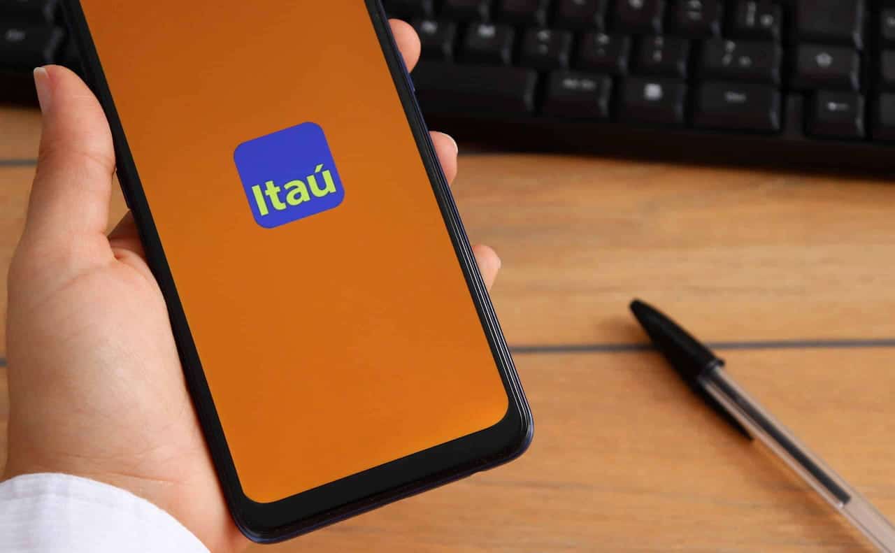 Mão segurando celular com app Itaú aberto em celular. Caneta e teclado de computador aparecem em segundo plano