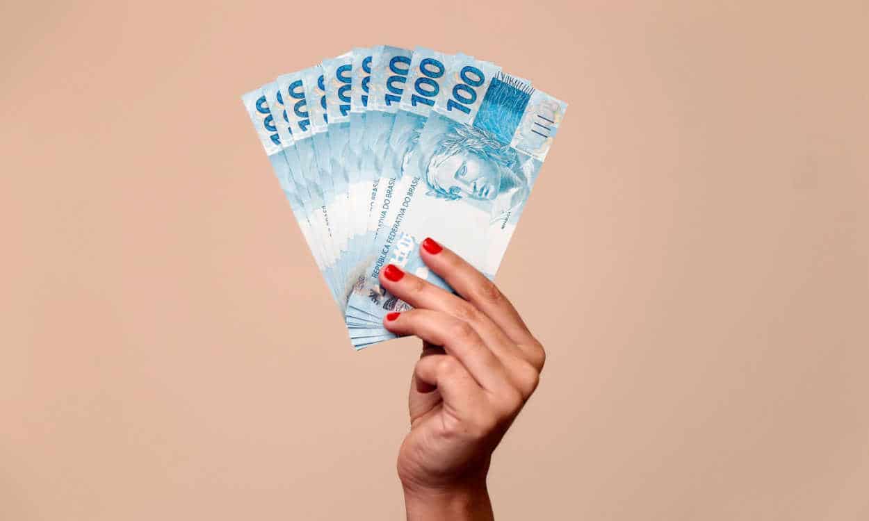 Mão segurando diversas notas de cem reais em leque. Fundo rosa. INSS Justiça federal RPVs